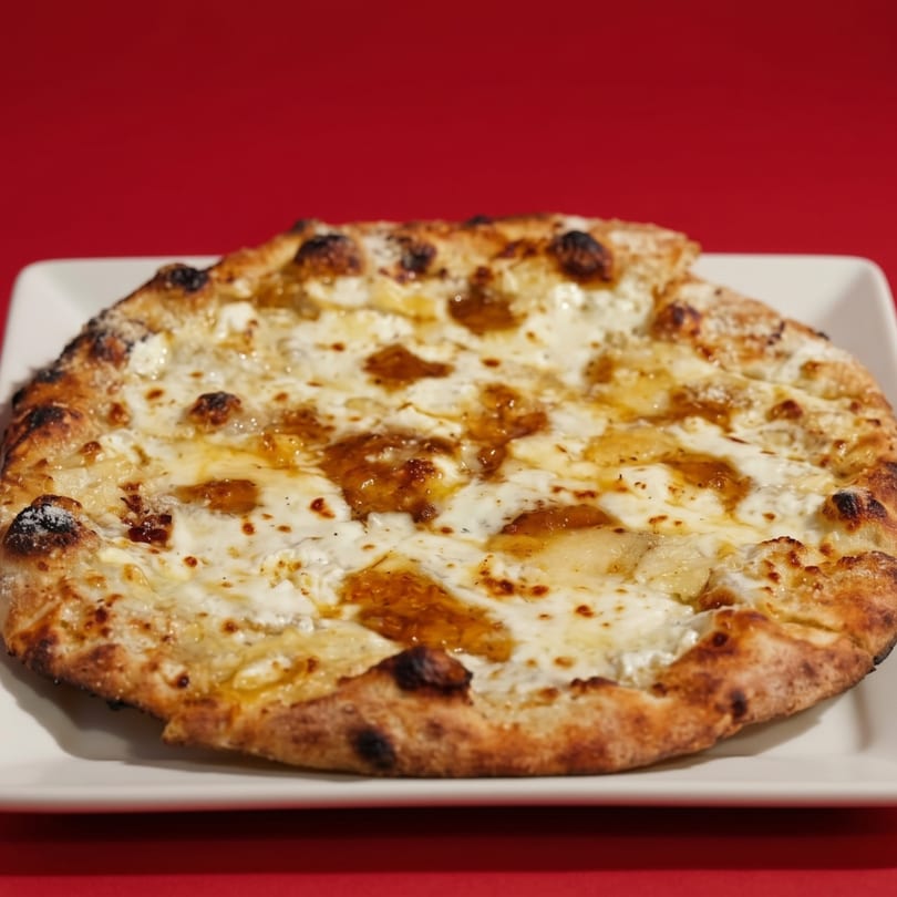 A white cheese pizza with dollops of golden honey on a white plate against a red background.